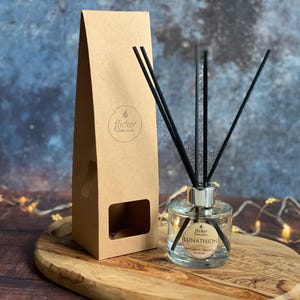 Puede incluir: Un difusor de aroma de vidrio transparente con cañas negras y una caja de papel marrón. La botella tiene una tapa plateada y una etiqueta que dice "LUNATHION" con un diseño floral. La caja tiene un logotipo que dice "flicker candle studio".