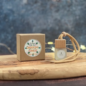 Puede incluir: Una pequeña botella de vidrio transparente con una tapa de madera y una cuerda beige, junto a una caja de cartón marrón. La caja tiene una pegatina con el texto "Cindersap Forest Maple Syrup". El fondo es borroso, azul oscuro con pequeñas luces.