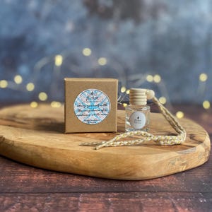 Könnte beinhalten: Eine kleine, klare Glasflasche mit Holzverschluss und geflochtener Kordel, neben einer kleinen braunen Schachtel. Die Schachtel und die Flasche stehen auf einem Holzservierbrett. Die Schachtel hat ein rundes Etikett mit dem Text "Flicker Candle Co."