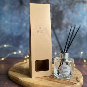 Puede incluir: Un difusor de aceites esenciales de vidrio transparente con cañas negras y una tapa plateada, junto a una caja de cartón marrón alta. La caja tiene un logotipo circular y una ventana rectangular. El difusor está sobre una tabla de madera.