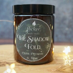 May include: A brown glass candle jar with a black lid and a black and white label that reads "flicker candle studio. THE SHADOW FOLD. CLOVE, PATCHOULI & MOSS"