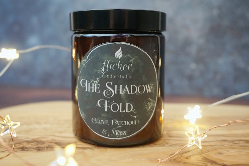 May include: A brown glass candle jar with a black lid and a black and white label that reads "flicker candle studio. THE SHADOW FOLD. CLOVE, PATCHOULI & MOSS"