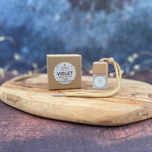 Może przedstawiać: Małe brązowe pudełko kartonowe z białą etykietą, na której widnieje napis "Flicker  Handcrafted  VIOLET  I WILL NOT DIE TODAY" oraz mała szklana butelka z drewnianym korkiem i przymocowanym beżowym sznurkiem. Butelka i pudełko leżą na drewnianej desce do krojenia.