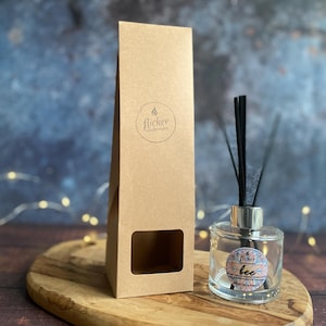May include: A clear glass diffuser bottle with black reeds and a silver cap, next to a brown paper box. The bottle has a label that says "bee". The box has a circular logo and the words "flicker candle co."
