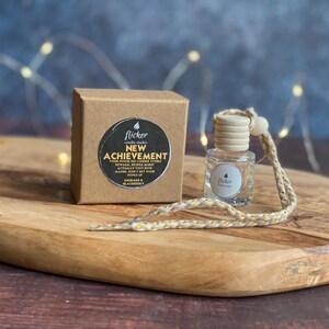 May include: A brown cardboard box and a small glass bottle with a wooden top are on a wooden tray. The box has a black and white sticker that reads "NEW ACHIEVEMENT". The bottle has a string attached. The background is a blurred blue.