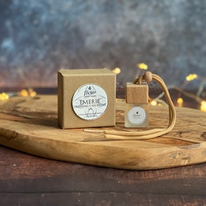 Könnte beinhalten: Eine braune Pappbox mit einem weißen Kreis, der den Text "Flicker Candle Studio" und "Emerie Nothing Can Break" enthält, mit den Worten "Cinnamon Honey & Clove" darunter. Ein hölzerner Auto-Lufterfrischer mit einer braunen Schnur und einer durchsichtigen Glasflasche mit dem Text "Flicker Candle Studio" auf dem Etikett.