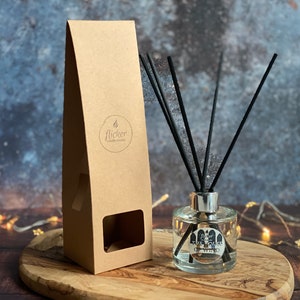 Könnte beinhalten: Ein Diffusor aus klarem Glas mit schwarzen Schilfrohren und einem silbernen Verschluss. Die Flasche trägt ein Etikett mit der Aufschrift "Flicker Candle Co. The House of the Wind" und steht auf einer Holzoberfläche mit einem braunen Papierkarton dahinter.