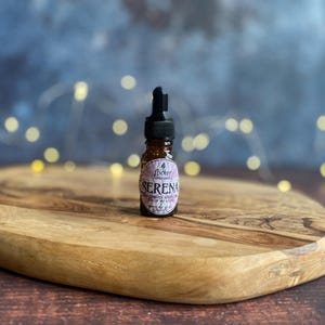 Könnte beinhalten: Kleine bernsteinfarbene Glasflasche mit schwarzem Tropfverschluss, beschriftet mit "Serena" in Weiß und Lila. Die Flasche steht auf einem Holzschneidebrett. Der Hintergrund ist ein verschwommenes Blau mit weichen, unscharfen gelben Lichtern.