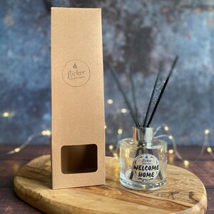 Könnte beinhalten: Eine klare Glas-Diffusorflasche mit schwarzen Schilfrohren und einem silbernen Deckel, neben einer hohen, braunen Pappschachtel. Die Flasche hat ein Etikett mit der Aufschrift "WELCOME HOME". Die Schachtel hat ein rundes Logo mit der Aufschrift "flicker candle studio".
