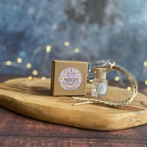Peut inclure: Une petite bouteille en verre avec un bouchon en bois et une corde tressée, à côté d'une petite boîte marron. La boîte porte une étiquette circulaire avec le mot "MISERY" et d'autres textes. Les articles sont sur une planche à découper en bois.
