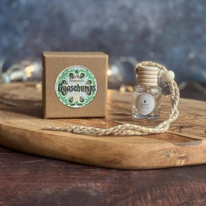 May include: A clear glass bottle with a wooden cap and a woven cord, alongside a brown cardboard box. The box features a circular sticker with the text "Goosebumps" and a green and white design. The bottle is for car air freshener.