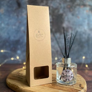 Könnte beinhalten: Ein klarer Glas-Diffusor für ätherische Öle mit schwarzen Schilfrohren und einer silbernen Kappe steht auf einer Holzoberfläche neben einer braunen Papierbox. Die Box hat ein Fenster und ein rundes Logo mit der Aufschrift "flicker candle studio". Der Diffusor hat ein Etikett mit der Aufschrift "zelsie".