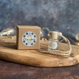 May include: A small, clear glass bottle with a wooden cap and a woven string, next to a brown cardboard box. The box has a circular label with the text "Coraline" and a button design. The items are on a wooden board.