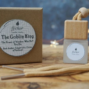 May include: A brown cardboard box with a white circle sticker that says "flicker candle studio. The Goblin King The Power of Voodoo. Who Do? You Do. Dark Amber, Ginger Lily, Cocoa & Patchouli" and a small glass bottle with a wooden top and a tan cord attached to it. The bottle has a white circle sticker that says "flicker candle studio."