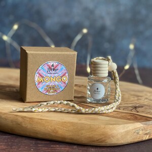 May include: A clear glass car air freshener with a wooden cap and a braided cord, next to a brown cardboard box. The box has a colorful sticker that reads "MONGO" and "Grape & Blueberry Slush". The items are on a wooden cutting board.