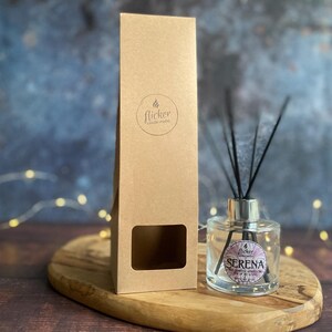 Könnte beinhalten: Eine transparente Glasdiffusorflasche mit schwarzen Schilfrohren und dem Wort "Serena" auf dem Etikett. Eine braune Pappschachtel mit dem Logo "flicker candle studio" befindet sich neben dem Diffusor auf einem Holztablett.