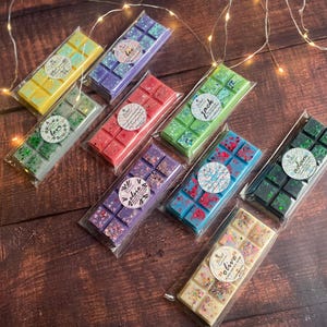 May include: An assortment of colorful wax melts in clear packaging, arranged on a dark wood surface. The melts are various colors, including yellow, purple, red, green, blue, and cream. Each melt has a decorative label with a name. Fairy lights are scattered around the melts.