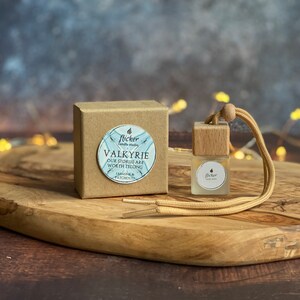 Könnte beinhalten: Eine kleine braune Schachtel mit einem weißen Aufkleber, auf dem "Valkyrie Our Stories Are Worth Telling Jasmine & Patchouli" steht, und eine kleine Glasflasche mit einem Holzdeckel und einer braunen Schnur daran. Die Flasche ist mit einer klaren Flüssigkeit gefüllt und hat einen weißen Aufkleber mit der Aufschrift "Flicker Candle Studio".