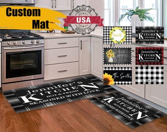 May include: Two black and white kitchen rugs with the text "Jennifer's Kitchen Hot & Fresh Seasoned with Love" in white. The rugs are on a wood floor in a kitchen.