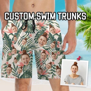 May include: Custom swim trunks with a tropical floral pattern and faces. The trunks feature a white background with green leaves, pink flowers, and multiple faces. The text "CUSTOM SWIM TRUNKS" is displayed at the top.
