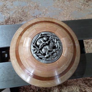 May include: A circular wooden decorative item with a central metal medallion depicting two koalas. The wood features light and reddish-brown tones, arranged in a circular pattern. The medallion is framed by a detailed leaf design.