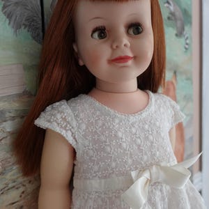 May include: A vintage doll with auburn hair and green eyes, wearing a white lace dress with a bow. The doll has a pearl-like headband. The background features a painted mural with woodland creatures.