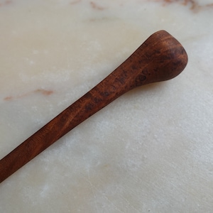 May include: A long, thin, wooden spoon with a rounded end. The spoon is dark brown and has a natural wood grain.