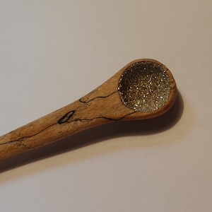 May include: A wooden spoon with a round, gold glitter-filled bowl. The spoon is made of light brown wood with dark brown streaks.