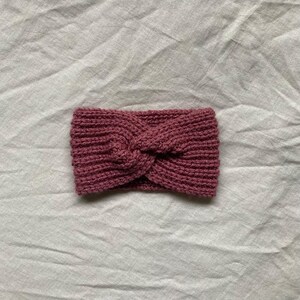 Könnte beinhalten: Ein gestricktes Stirnband in einem staubigen Rosaton. Das Stirnband hat ein verdrehtes Knotendesign in der Mitte. Das Material scheint ein weiches Garn zu sein, und das Stirnband liegt auf einer weißen, strukturierten Oberfläche.