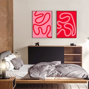 May include: Two abstract art prints with red lines on a pink and red background. The prints are framed in black and hung on a white wall above a wooden bed frame.