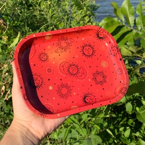 May include: A red metal rolling tray with a gold sun and moon pattern. The tray is decorated with a repeating pattern of suns, moons, and stars.