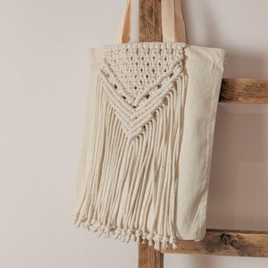 Puede incluir: Bolso tote de macramé color crema con diseño geométrico y detalles de flecos. El bolso cuelga de una escalera de madera rústica. El bolso tiene un cuerpo y correas de lona color crema.
