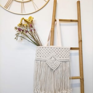 Puede incluir: Bolso tote de macramé color crema con diseño geométrico y largas borlas. La bolsa cuelga de una escalera de bambú, con flores secas en su interior. Un reloj dorado es visible en el fondo.