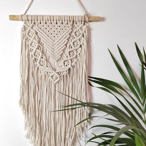 Puede incluir: Colgante de pared de macramé color crema con un diseño geométrico, suspendido de una varilla de madera. El intrincado anudado crea un patrón texturizado, con largas franjas en la parte inferior. Una pieza decorativa para la decoración del hogar.