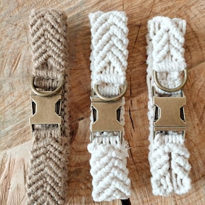 Puede incluir: Tres collares para perros de macramé en tonos naturales. Los collares presentan un patrón de espiga tejido y herrajes de latón antiguo. Uno es marrón claro y dos son blanquecinos. Los collares se exhiben sobre una superficie de madera.