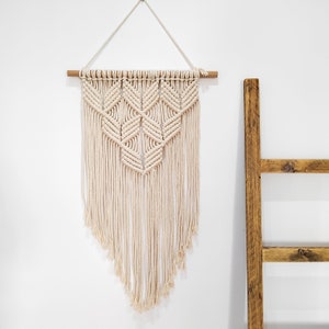 Puede incluir: Un tapiz de pared de macramé color crema con un diseño geométrico. La decoración de la pared está suspendida de un pasador de madera y presenta una forma triangular con extremos largos y con flecos. Una escalera de madera está en el fondo.