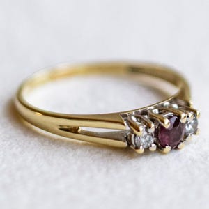 Può includere: Un anello in oro con una pietra preziosa rosso scuro a taglio rotondo al centro, affiancata da due pietre chiare più piccole a taglio rotondo. La fascia è realizzata in oro giallo lucido, con un design diviso che porta alle montature.