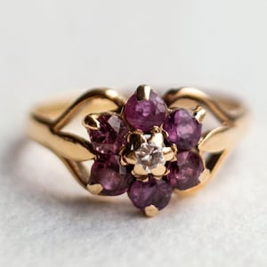 May include: A gold ring with a flower design featuring a cluster of purple gemstones and a single white gemstone in the center.
