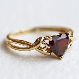 Op de afbeelding: Een gouden ring met een hartvormige, donkerrode edelsteen in het midden. De ringband heeft een gespleten ontwerp dat samenkomt in een decoratieve zetting die de steen vasthoudt. Het metaal heeft een gepolijste afwerking en de steen is gefacetteerd.