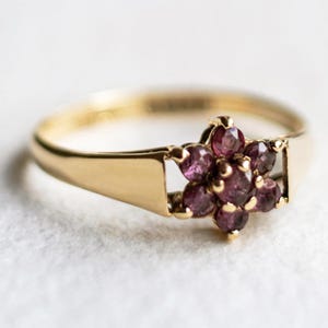 Peut inclure: Une bague en or ornée d'un groupe de sept pierres précieuses rondes d'un violet pourpre profond disposées en motif floral. L'anneau est lisse et poli, se connectant au sertissage central par une structure ouverte simple.