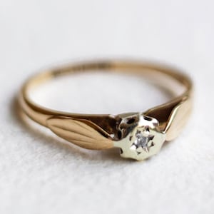 Peut inclure: Une bague en or dotée d'un sertissage central avec une petite pierre ronde transparente. L'anneau présente des motifs en forme de feuilles de chaque côté de la pierre, passant d'une finition en or poli à un métal argenté au niveau du sertissage.