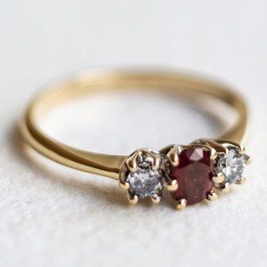 Peut inclure: Une bague en or ornée d'une pierre précieuse rouge foncé de taille ovale au centre, encadrée par deux pierres blanches transparentes de taille ronde. L'anneau est lisse et poli, avec les trois pierres maintenues par de petites griffes.