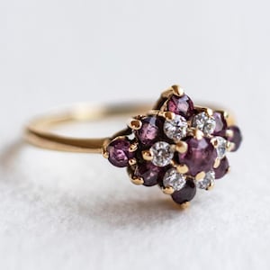 Può includere: Un anello in oro con una montatura a grappolo caratterizzata da una pietra preziosa rotonda viola centrale circondata da pietre più piccole, sia viola che bianche trasparenti. Il gambo dell'anello è sottile e in oro lucido.