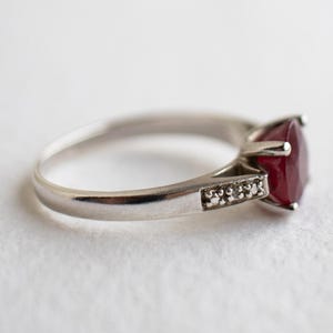 Op de afbeelding: Een zilverkleurige ring met een opvallende, ronde, dieprode edelsteen in een zetting met vier pootjes. De ringband is versierd met een klein rijtje piepkleine, heldere, fonkelende steentjes die aan de zijkant bij de zetting zijn ingelegd.