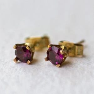 Peut inclure: Une paire de boucles d'oreilles à clous avec des pierres précieuses rondes de couleur magenta profond serties dans des griffes en métal doré. Chaque boucle possède un fermoir à tige, révélant une taille à facettes qui reflète la lumière.