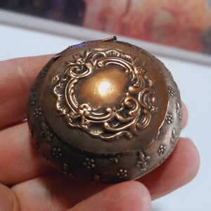 Könnte beinhalten: Eine kleine, runde, antike, messingfarbene Schmuckdose mit einem verzierten Blumenmuster. Der Deckel ist geschlossen und die Dose liegt auf einer Hand.