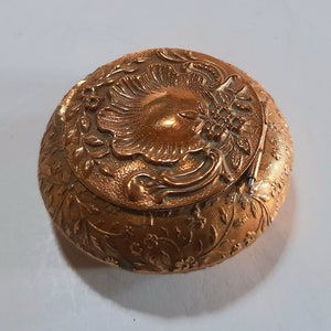 Può includere: Una piccola scatola rotonda in metallo dorato con un design floreale ornato. Il coperchio presenta un motivo floreale in rilievo dettagliato.