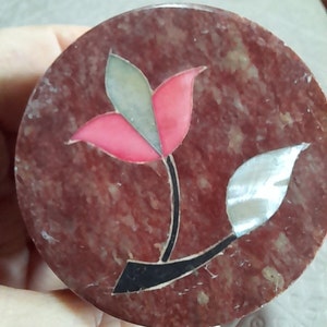 Könnte beinhalten: Eine runde, rot-braun-weiße Marmor-Schmuckdose mit einem Blumendesign. Die Blume hat rosa und grüne Blütenblätter und einen schwarzen Stiel mit einem weißen Blatt.