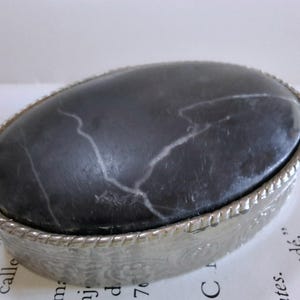 May include: A small, oval-shaped trinket box with a silver-coloured metal rim and a black marble top. The marble has white veins running through it.