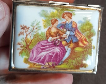 May include: A small, rectangular, silver-colored box with a hinged lid. The box is decorated with a porcelain panel depicting a couple in a romantic setting. The couple is dressed in 18th-century clothing and is surrounded by trees and flowers.